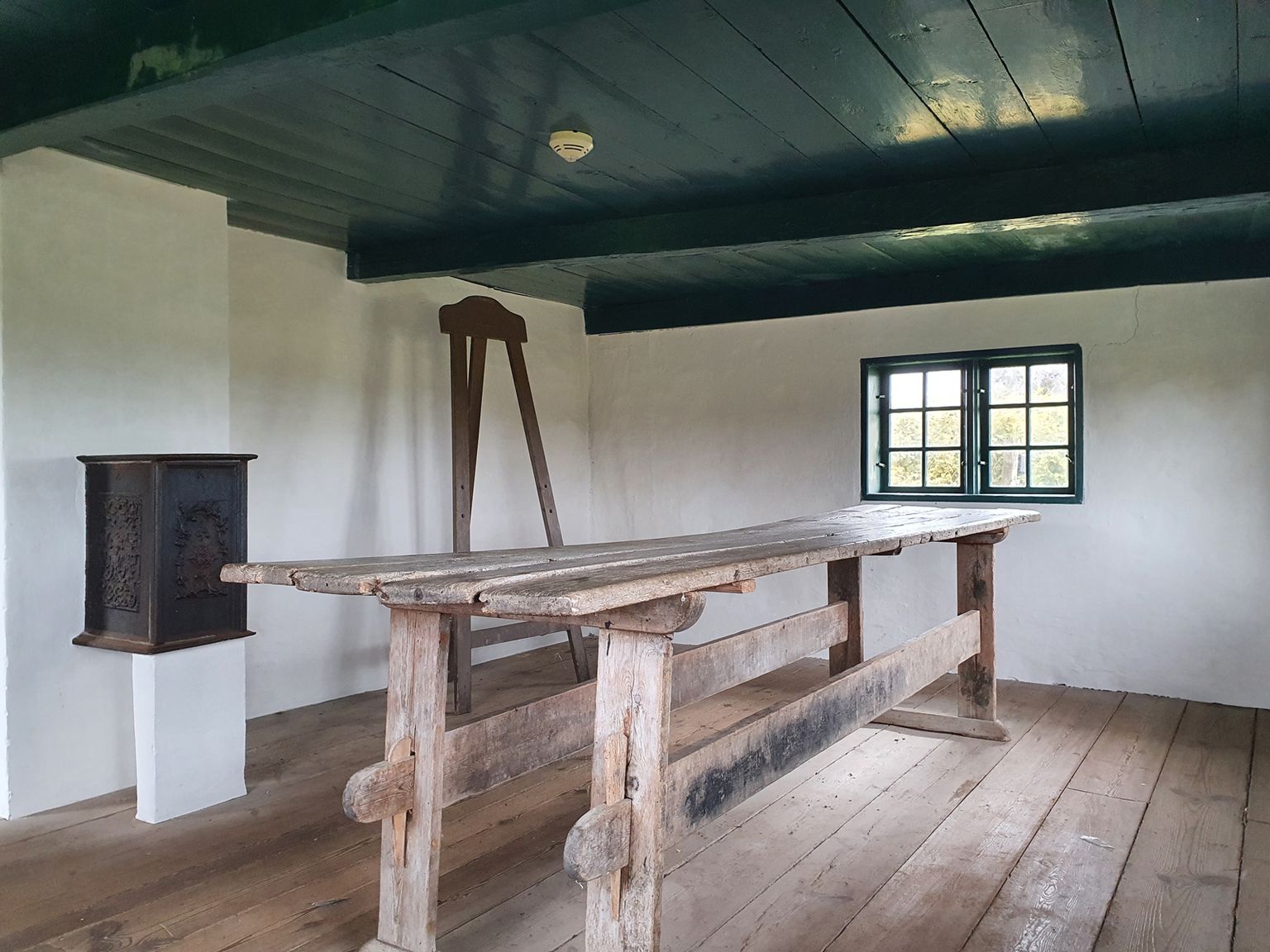 Interiør i Toftum Skole med originalt langbord, stol og jernkakkelovn under det grønmalede bjælkeloft