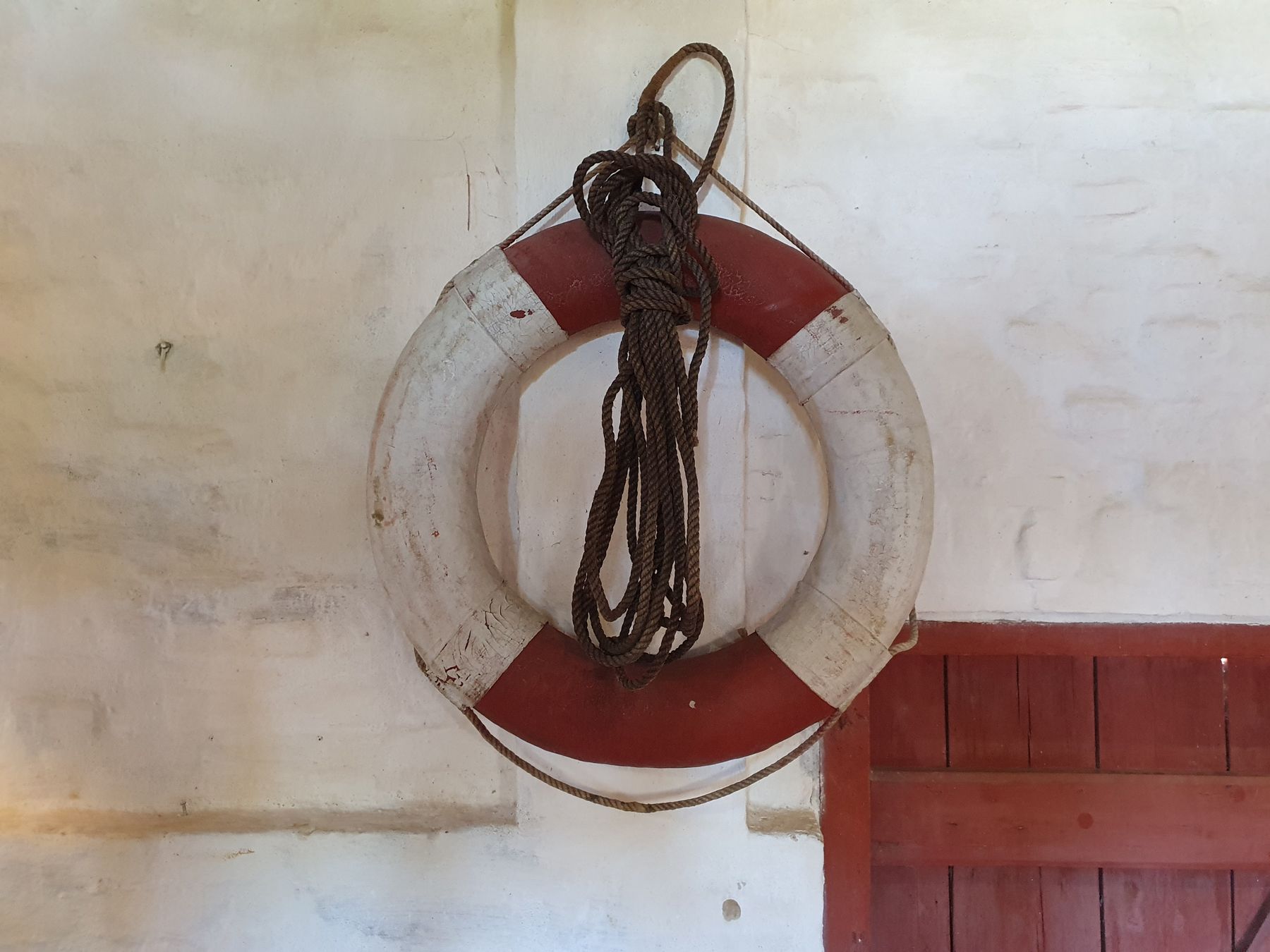 Historisk rød-hvid redningskrans med reb ophængt på hvidkalket væg i udstillingen om strandinger og forlis