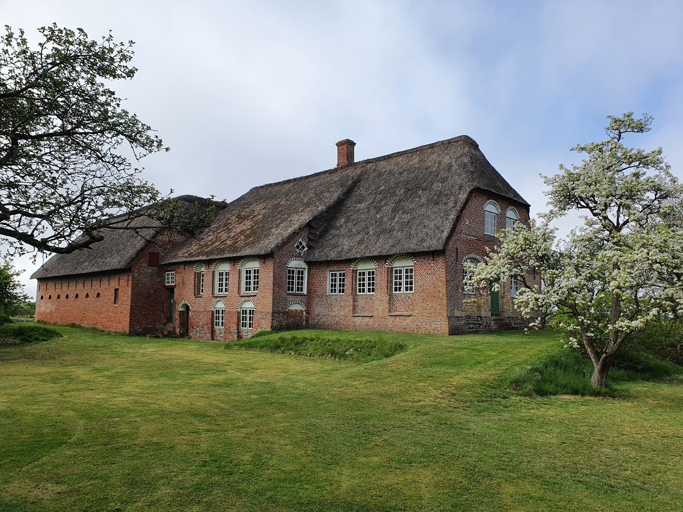 Kommandørgårdens hovedbygning set fra haven med stråtag, røde mursten, bueformede vinduer og blomstrende frugttræer