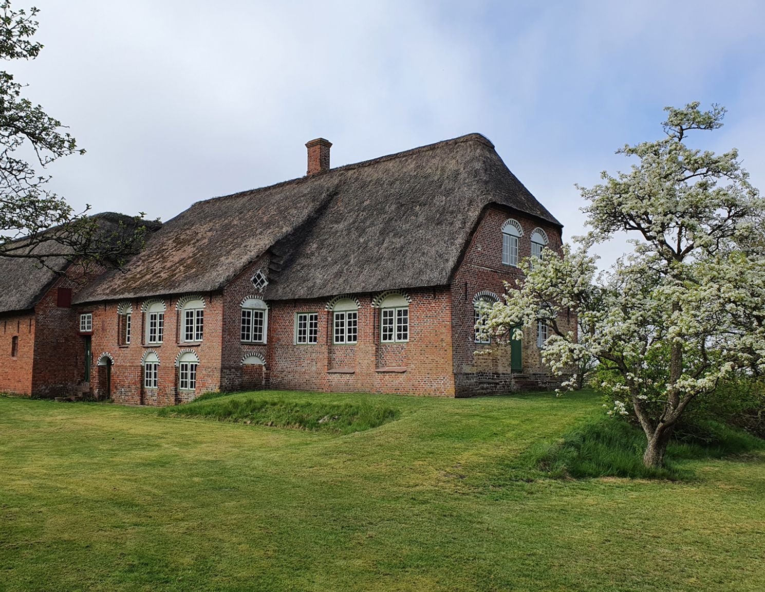The Sea Captain's House main building seen from the garden with thatched roof, red brick, arched windows, and blossoming fruit trees