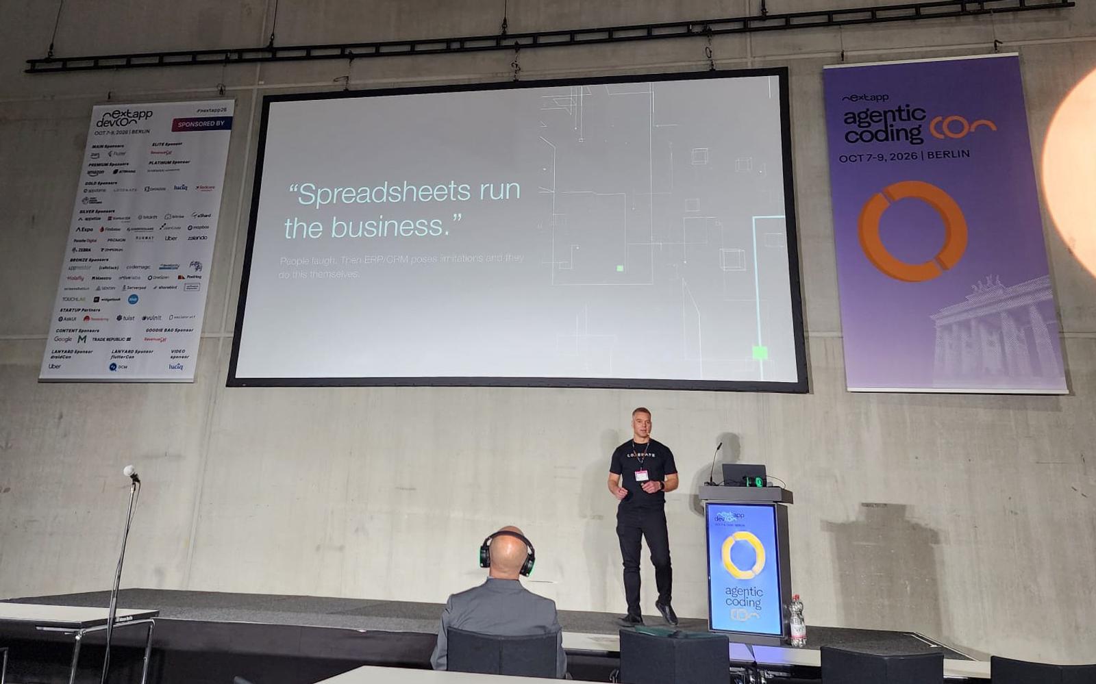 Ville Lindfors on stage at Next.app DevCon 2026 in Berlin, with a slide reading "Spreadsheets run the business."