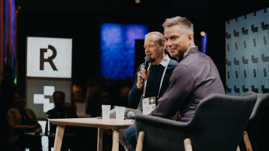 Two men speaking on stage during a panel discussion, one holding a microphone and the other smiling toward the camera.