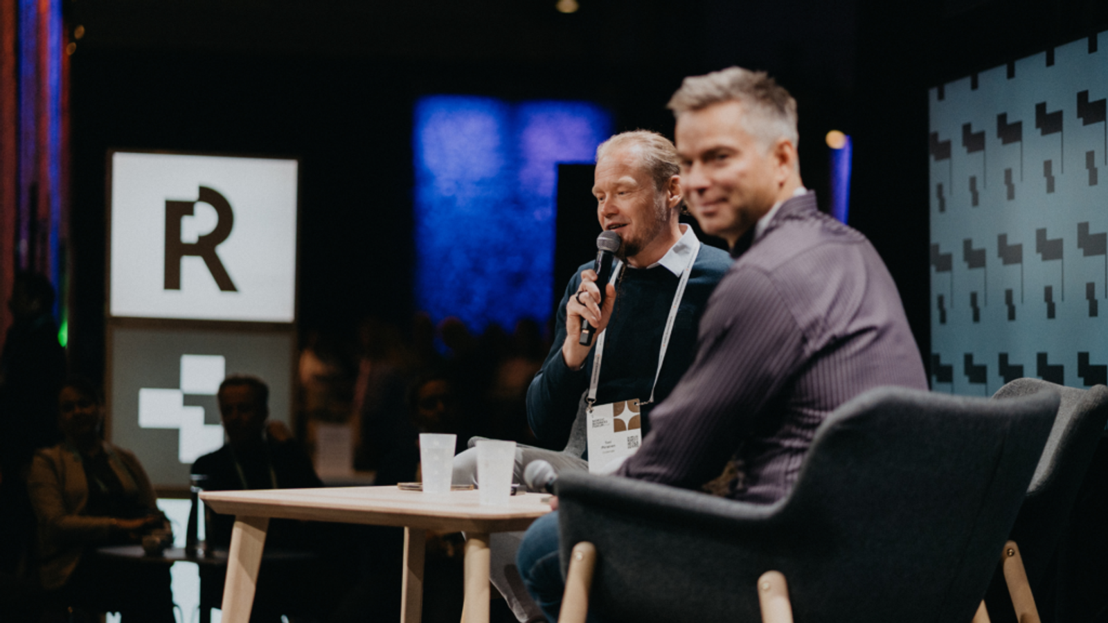 Two men speaking on stage during a panel discussion, one holding a microphone and the other smiling toward the camera.