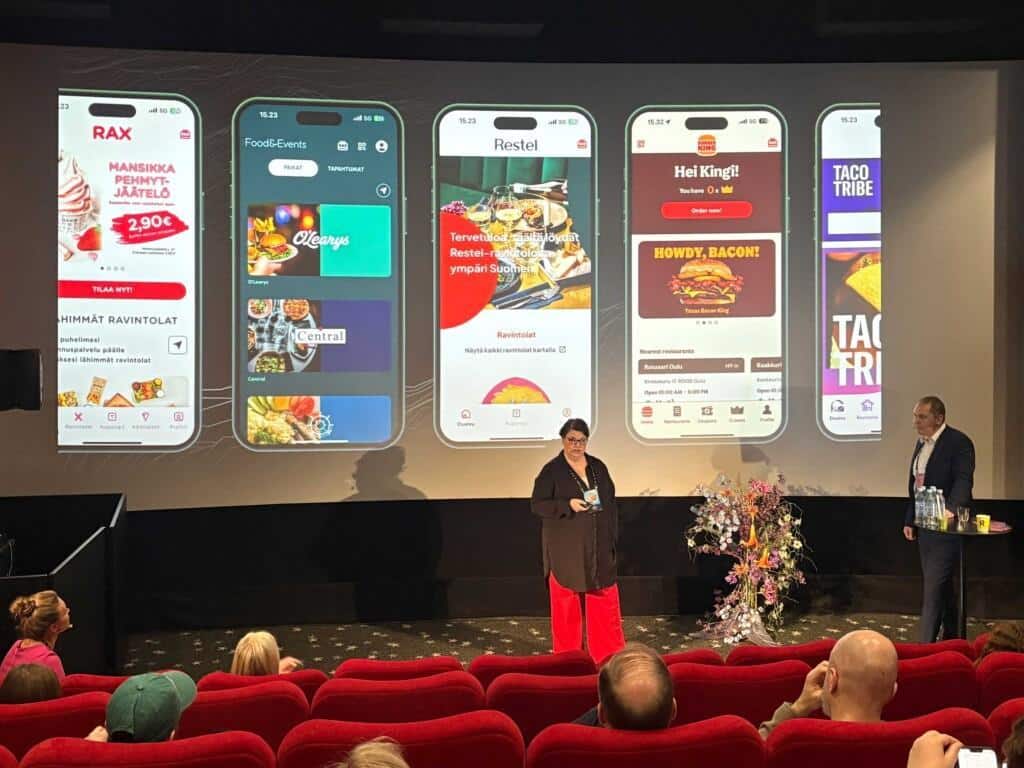 A presentation slide displaying five mobile app screens for restaurant brands including RAX, Food&Events, Restel, Burger King, and Taco Tribe. Two speakers stand in front of the screen, engaging with the audience seated in red cinema chairs.