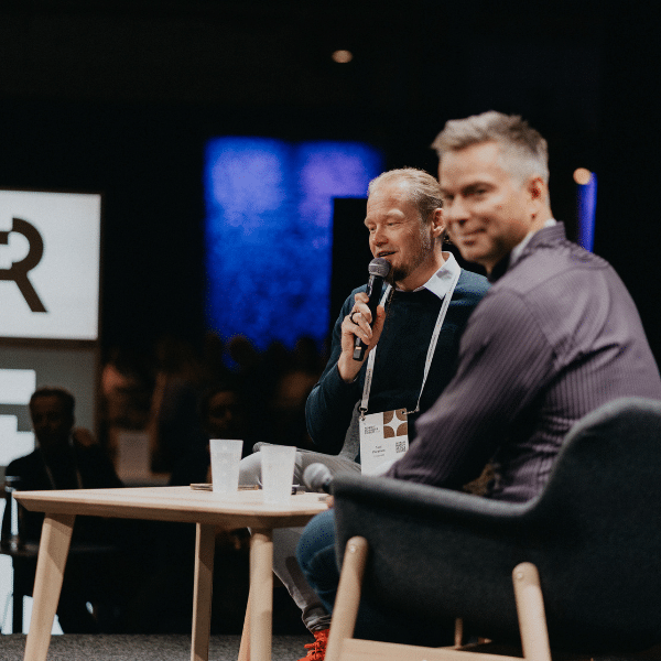 Two men speaking on stage during a panel discussion, one holding a microphone and the other smiling toward the camera.