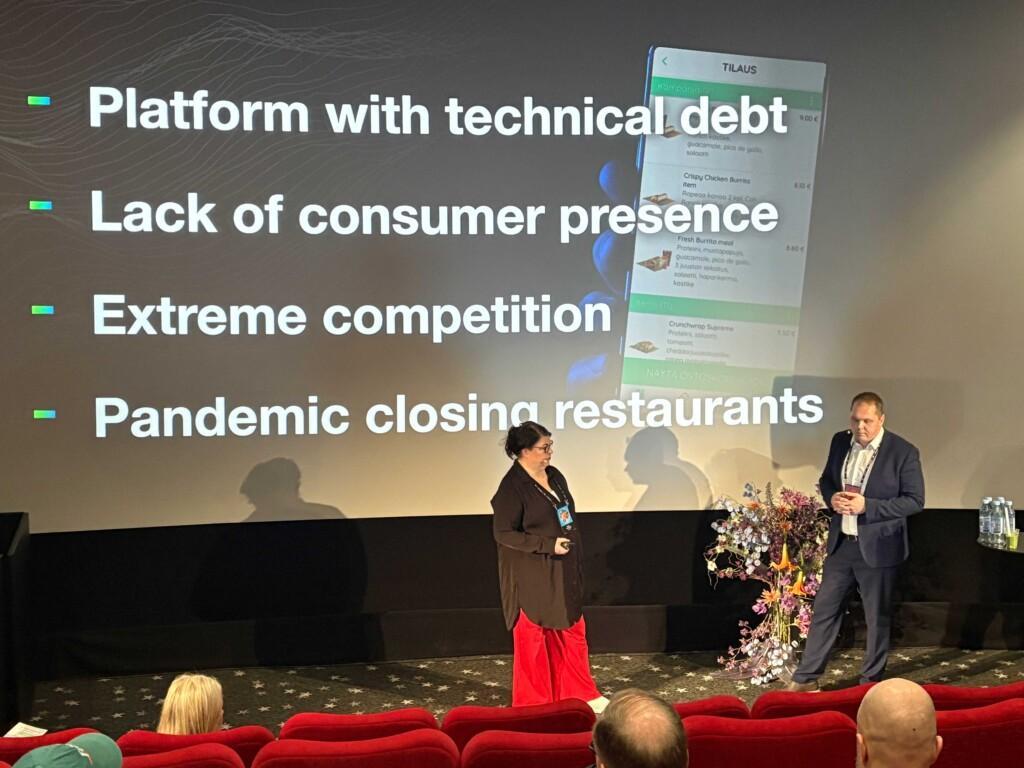 Two presenters speak on stage in front of a large screen showing a slide with business challenges: “Platform with technical debt, Lack of consumer presence, Extreme competition, Pandemic closing restaurants.” A smartphone app interface is partially visible in the background.