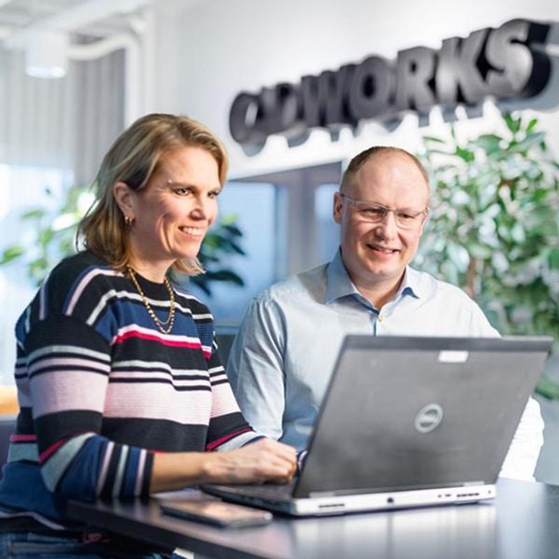 A woman and a man collaborating on a laptop in a modern office with "CADWORKS" branding on the wall.