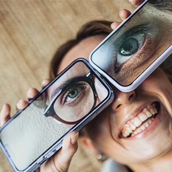 Hymyilevä henkilö pitelee kahta älypuhelinta kasvojensa edessä, joissa näkyy suurennettu kuva silmästä, havainnollistaen näkemistä ja visuaalista tarkastelua.