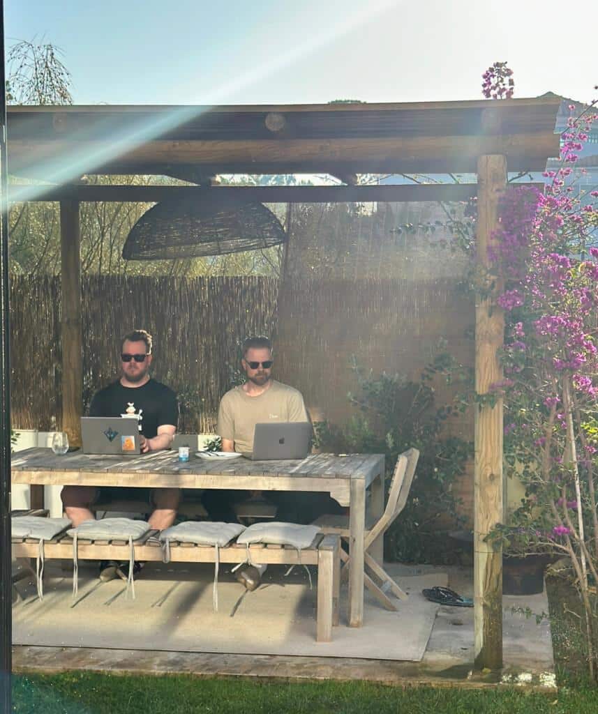 Mikael Komu ja Miika Toljamo Portugalissa etätöissä. He istuvat puisen pöydän ääressä, kasvot kohti kameraa kannettavat tietokoneet edessään.