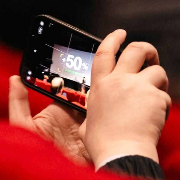 A close-up of a person’s hands holding a smartphone horizontally, capturing a photo of a presentation slide. The slide on the screen displays “+50%” in large text with an audience seated in red cinema chairs.