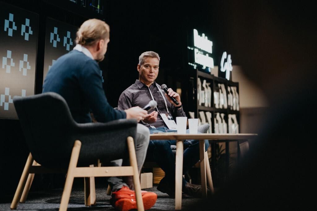 A closer view of the same two speakers on stage at the Reaktor Ecosystem Café during Nordic Business Forum. One is speaking into a microphone while the other looks toward the audience. Cups and notes are on the small table between them.