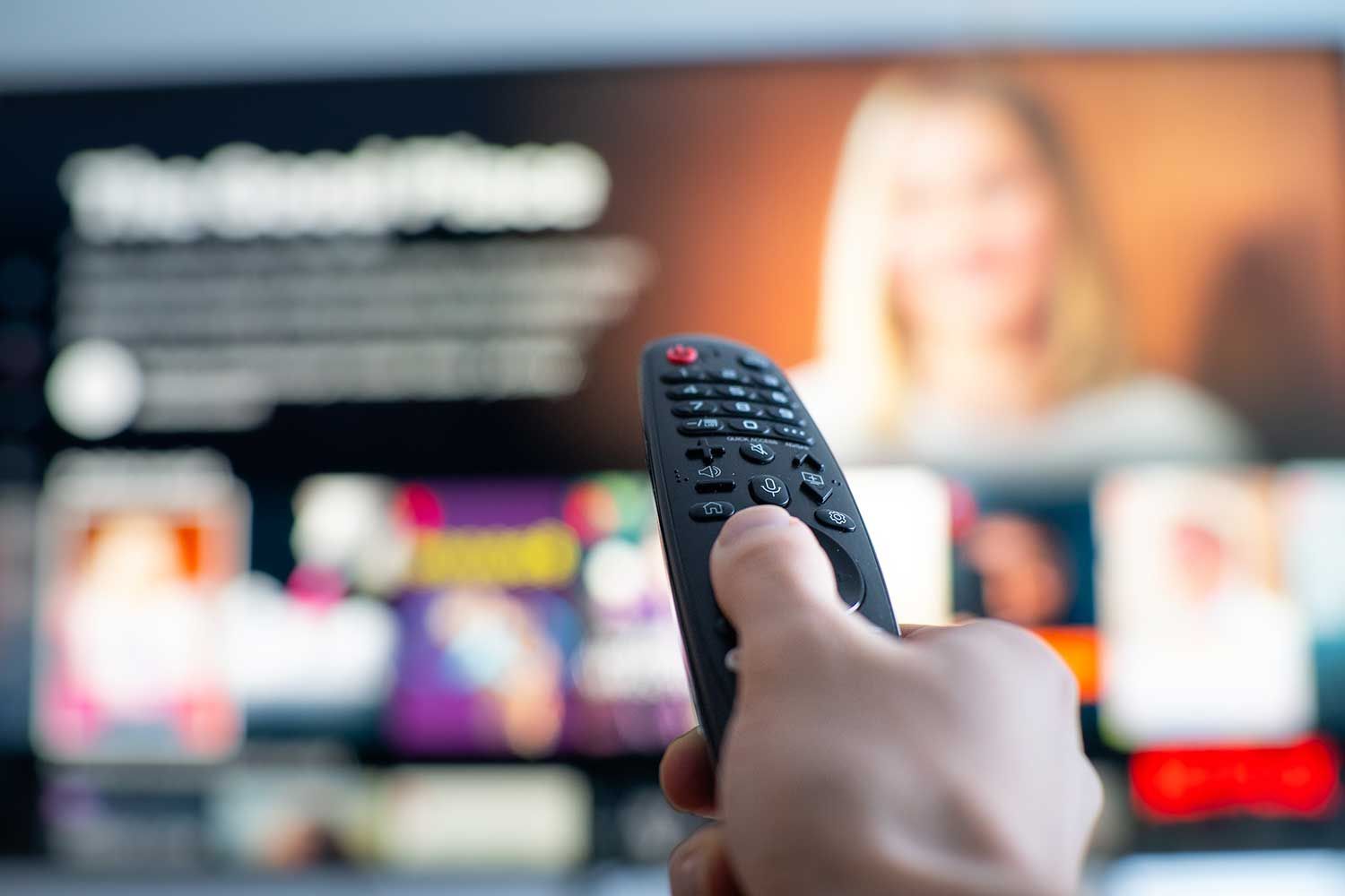 A person holding a TV remote control and pointing it towards a blurred television screen displaying a streaming service interface with multiple show thumbnails and a programme titled "The Good Place".