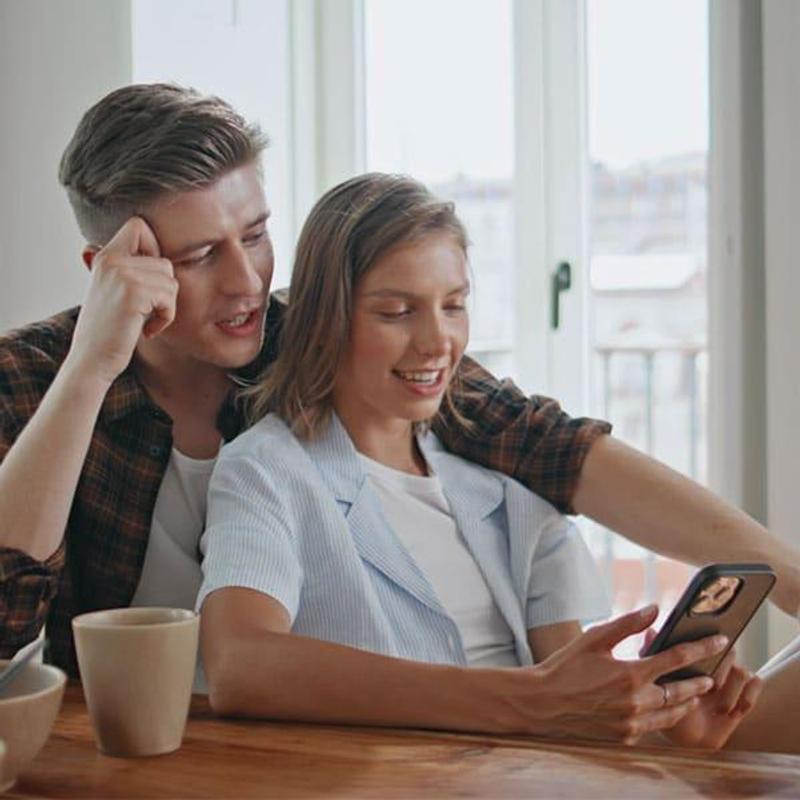 Young couple sitting at their kitchen table, smiling and browsing a smartphone together, suggesting they are looking for a new home or listing online.