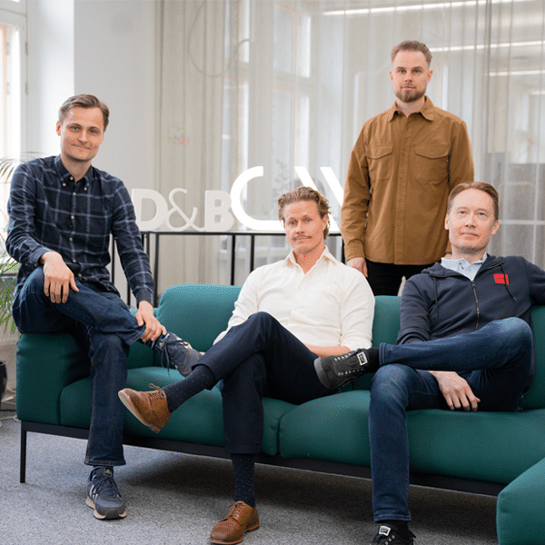 Four men pose for a professional group photo in a modern office setting. Three are seated on a green sofa, while one stands behind them. They are dressed in casual business attire, with a backdrop of large windows, a green potted plant, and a sign reading "D&BW." The mood is relaxed and confident, reflecting a collaborative team environment.