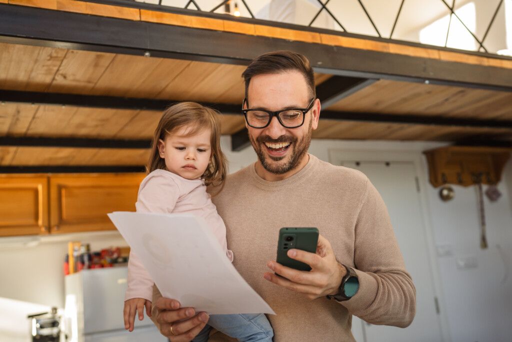 Aikuinen mies, jolla on parta ja silmälasit, pitää lasta sylissään kotikeittiössä. Hän hymyilee katsoessaan puhelintaan ja pitelee samalla paperia, jota lapsi tarkastelee vakavana. Taustalla näkyy puuhyllyjä ja parvirakennelma.