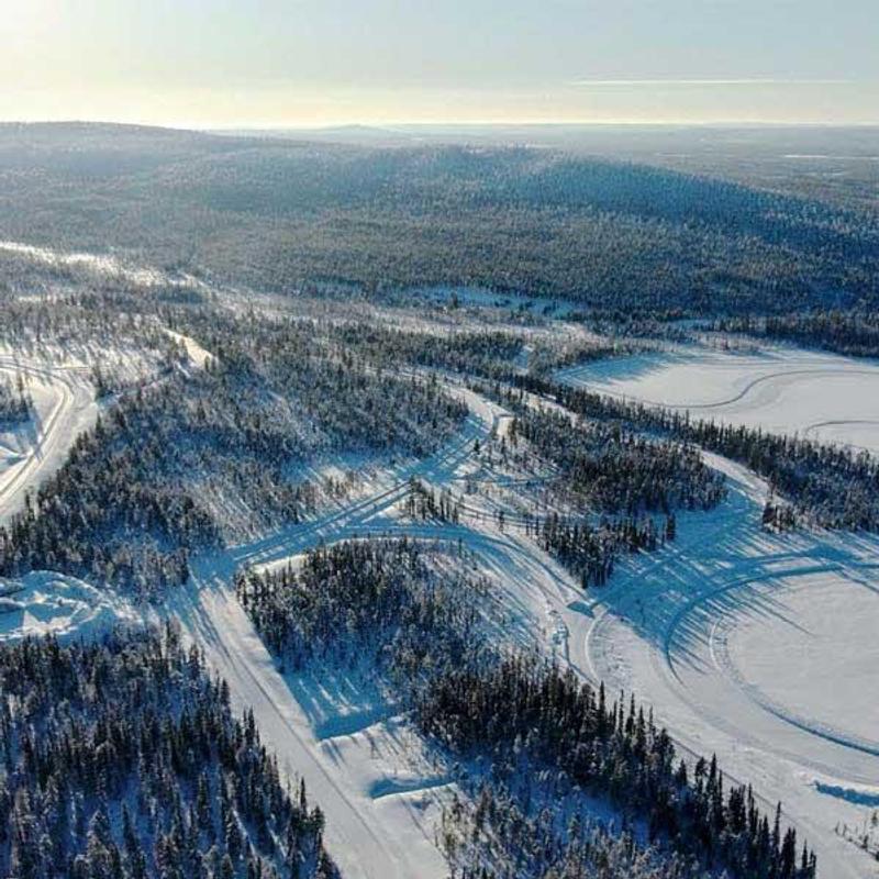 Drone view of a snowy proving ground with a variety of test track shapes, including a prominent circular track. The tracks are carved into the snow across a large open area, surrounded by dense forest, under a bright and crisp winter sky.