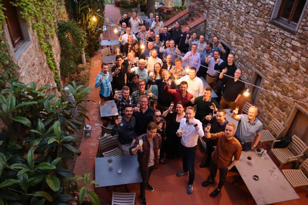 Group picture with Codemate people, they are smiling and raising glasses. There's stone walls and some greenery around them.