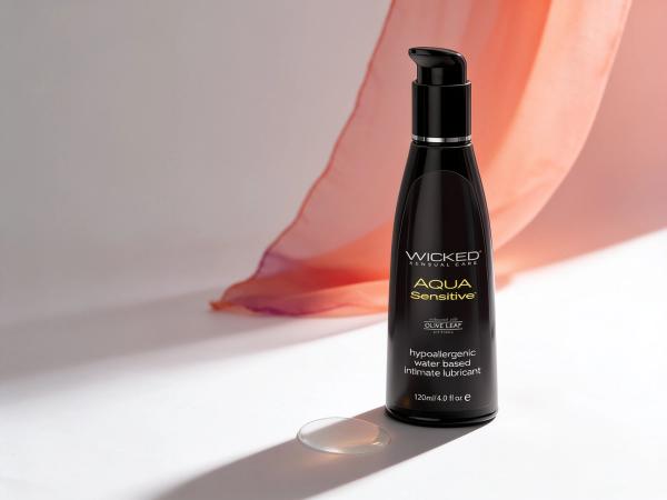 The Wicked Sensitive glycerin-free water-based lubricant bottle standing in warm daylight on white paper, a sheer coral fabric sweeping behind it and a single clear droplet at its base, illustrating this guide to choosing a lube for sensitive skin.