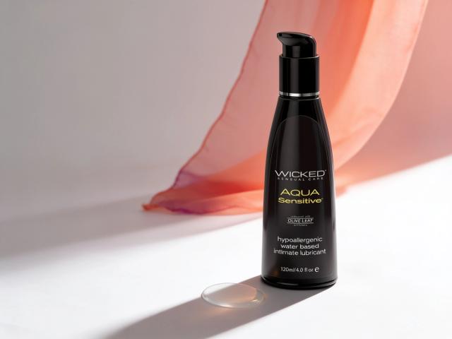 The Wicked Sensitive glycerin-free water-based lubricant bottle standing in warm daylight on white paper, a sheer coral fabric sweeping behind it and a single clear droplet at its base, illustrating this guide to choosing a lube for sensitive skin.