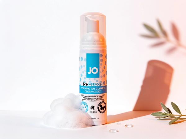 JO Refresh foaming toy cleaner bottle standing upright on a white surface in warm daylight, with a soft cloud of foam and clear water droplets beside it and a sage leaf sprig behind