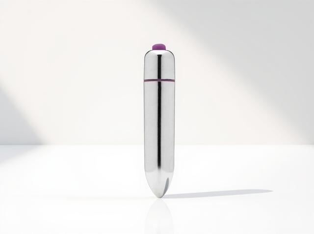 The 3-Speed Tantus Original Bullet Vibrator standing tall on a white surface in warm window daylight, its polished chrome body and purple accent ring catching a soft diagonal light and shadow.