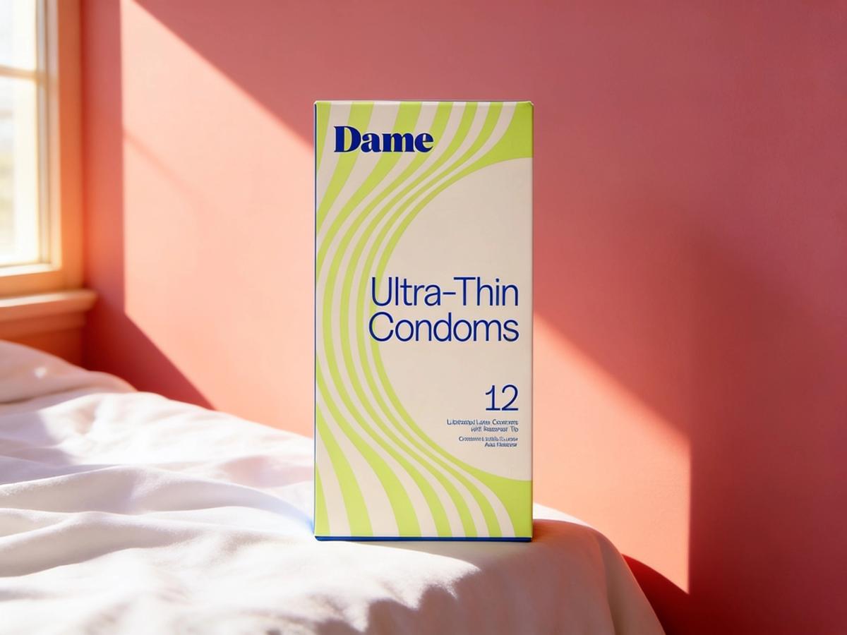 A box of Dame Ultra-Thin Natural Latex Condoms, 12-pack, standing on the edge of a sunlit white bed against a warm coral wall.