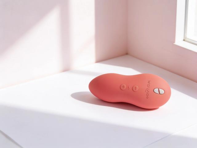 The We-Vibe Touch X in Crave Coral resting on white paper in a pale blush corner, soft window light tracing the matte silicone, its round buttons and the fine join along its edge