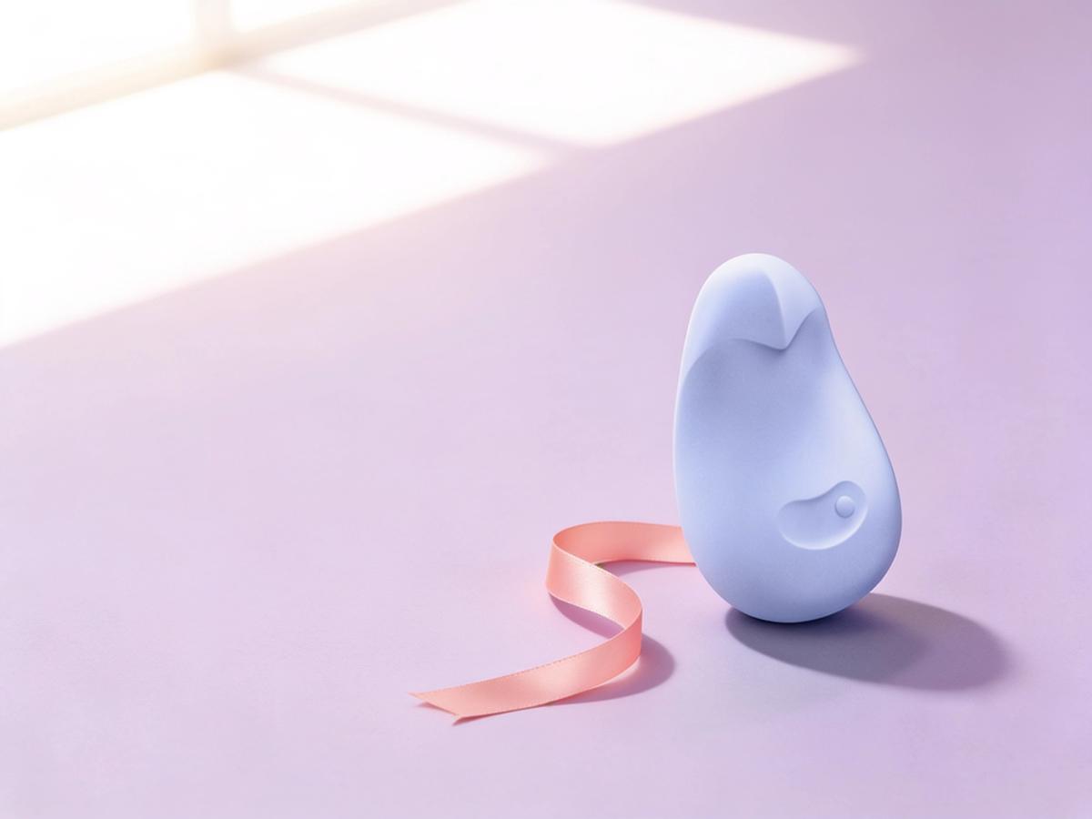The Dame Pom Flexible Palm Vibrator Ice standing upright in soft window light on a pale plum-tinted backdrop, a thin coral ribbon curling beside it