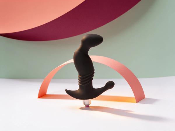 The Vibrating Prostate Play Onyx Massager standing upright on white paper, its curved matte black tip, ridged shaft and broad anchor arm all in full view, framed by a coral paper arch that echoes the curve, in warm daylight.