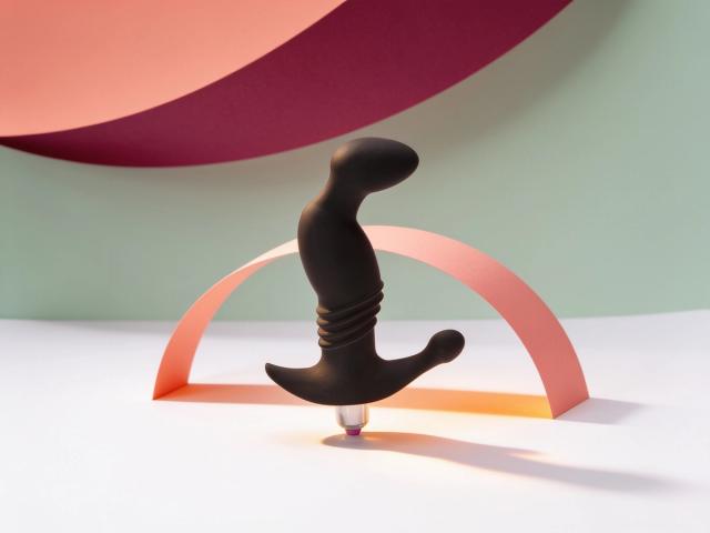 The Vibrating Prostate Play Onyx Massager standing upright on white paper, its curved matte black tip, ridged shaft and broad anchor arm all in full view, framed by a coral paper arch that echoes the curve, in warm daylight.