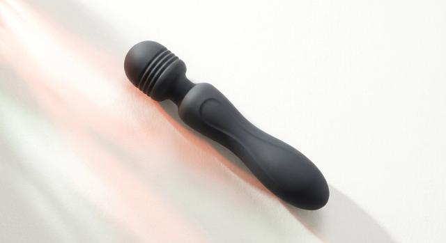 A black Domi 2 wand vibrator lying at an angle on a bright white surface, warm coral light streaming diagonally across the frame, its ribbed head and slim curved handle shown at true scale, illustrating the mid-size tier between compact bullets and full-size wands.