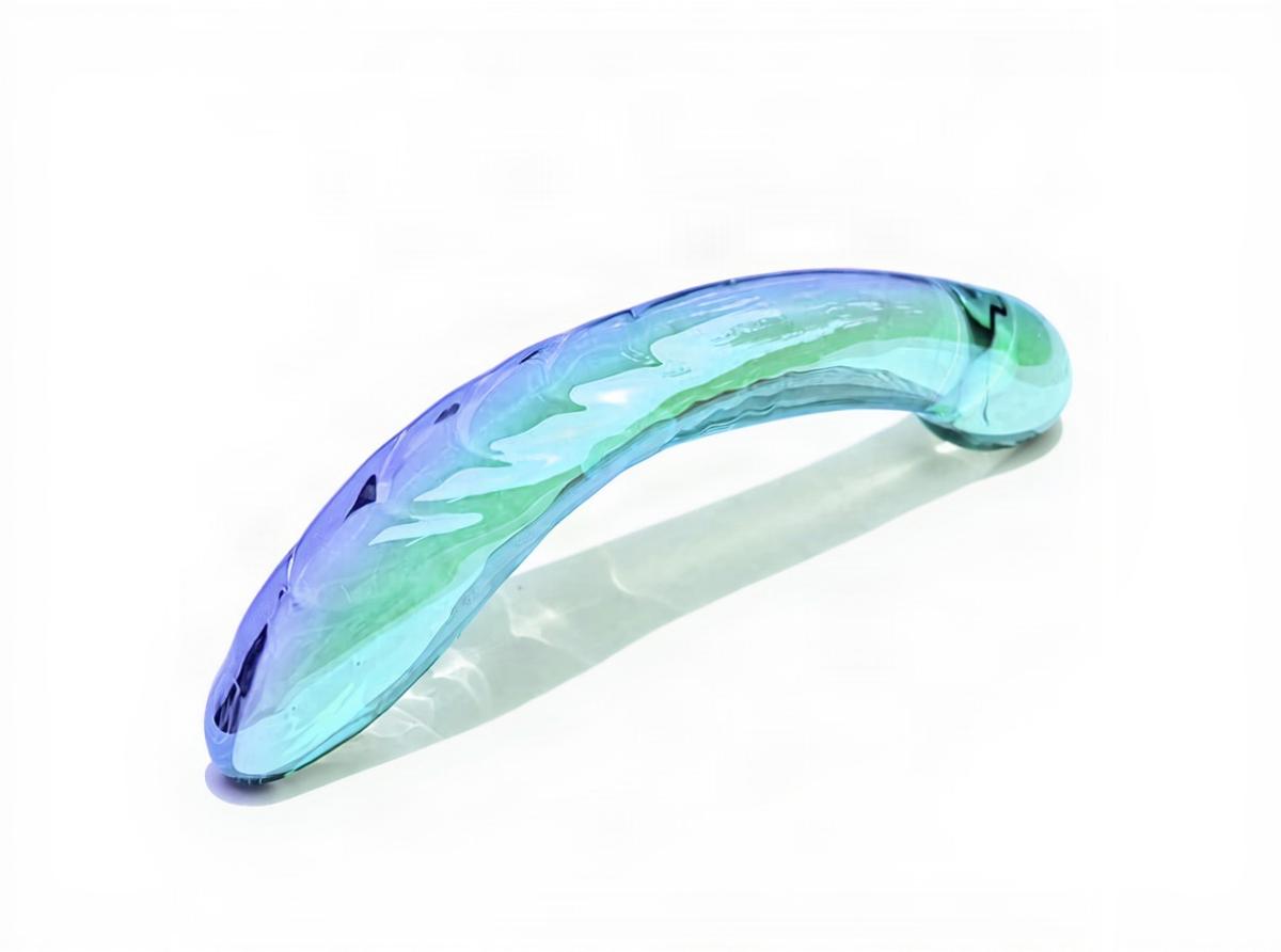 The Biird Kalii glass G-spot dildo, a curved borosilicate piece with an iridescent blue-violet to seafoam-green finish, resting on a bright white daylight surface, its smooth glossy surface catching clean highlights to show the non-porous material up close.