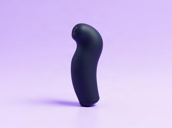 The Tantus Acute curved dildo in its deep night-sky silicone finish standing bold and upright on a bright plum-soft studio backdrop, its precise curve catching soft daylight, one half of the dildo-versus-vibrator comparison