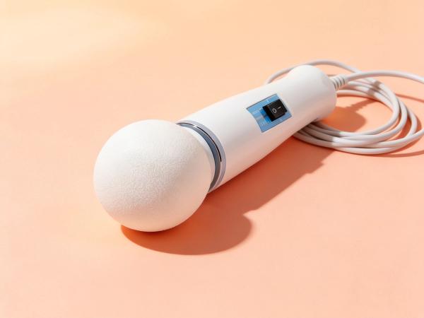 Magic Wand Original corded wand massager resting diagonally on a warm coral-soft surface, cord coiled neatly beside it, bright daylight editorial still life