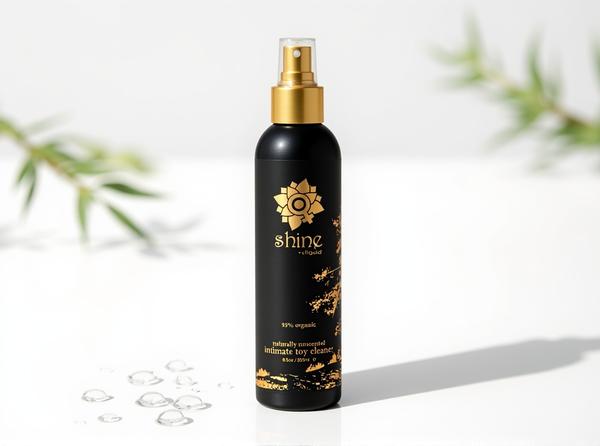 Sliquid Shine Organic Intimate Toy Cleaner spray bottle standing upright in bright daylight on a white surface, with scattered water droplets and a blurred sage-green leaf sprig suggesting a freshly rinsed vibrator left to air-dry.