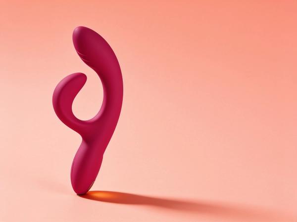 The We-Vibe Nova 2 flexible silicone rabbit vibrator leaning at a dynamic angle on a bright coral-soft backdrop, its curved outer arm and ribbed inner shaft both clearly visible to show how the flex lines up.