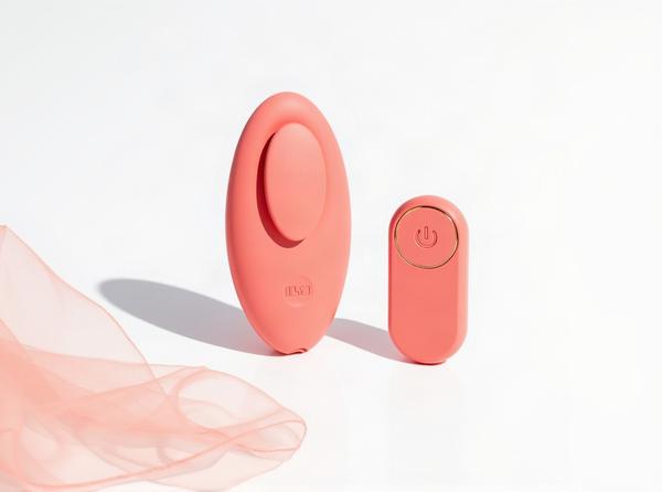 The ILY by Je Joue Panty Vibrator, a coral-pink wearable panty vibe, standing upright beside its round remote control on white, a sheer blush satin edge draped beneath it, staged in warm daylight the moment before it's worn.
