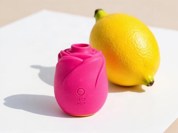 The ROMP Rose 2.0 Pleasure Air clitoral stimulator in bright fuchsia silicone, standing on white paper in warm daylight with a whole lemon resting behind it so the two shapes rhyme.
