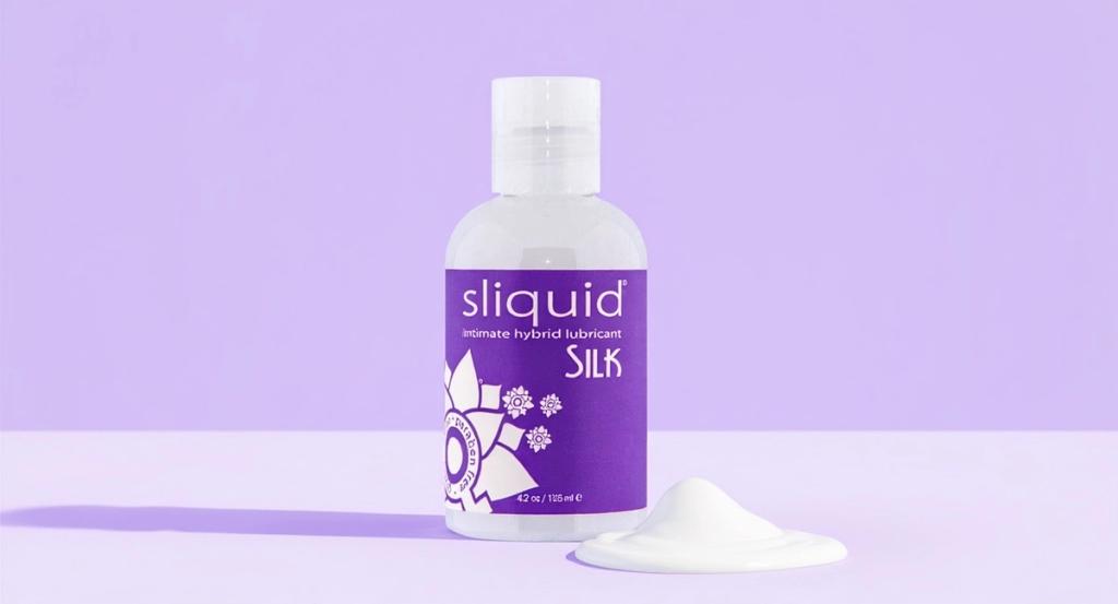 A single clear plum-labeled hybrid lubricant bottle stands large on a pale lilac studio ground with a soft sheen of glide catching bright daylight.