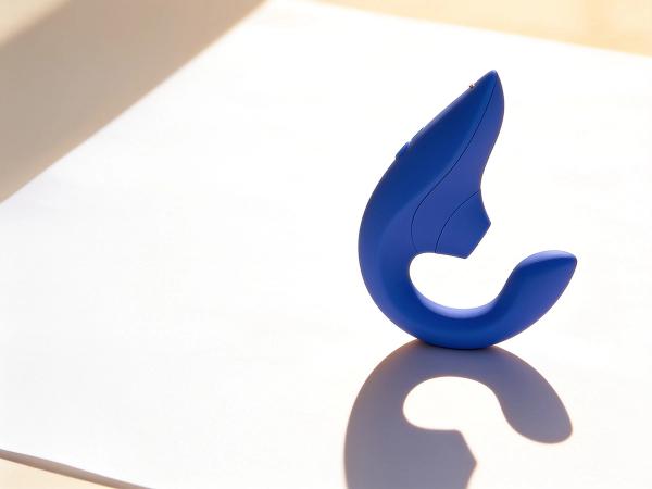 The vibrant blue Womanizer Blend dual-stimulation vibrator standing upright on a sunlit white paper surface, its curved two-arm silhouette clearly visible.
