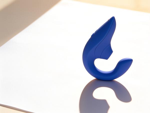The vibrant blue Womanizer Blend dual-stimulation vibrator standing upright on a sunlit white paper surface, its curved two-arm silhouette clearly visible.