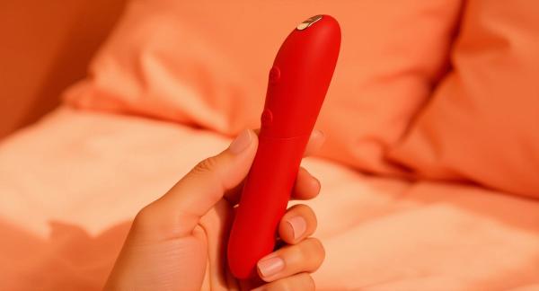 A cherry-red We-Vibe Tango X bullet vibrator held in a hand against warm coral bedside linens.