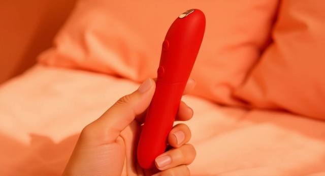 A cherry-red We-Vibe Tango X bullet vibrator held in a hand against warm coral bedside linens.