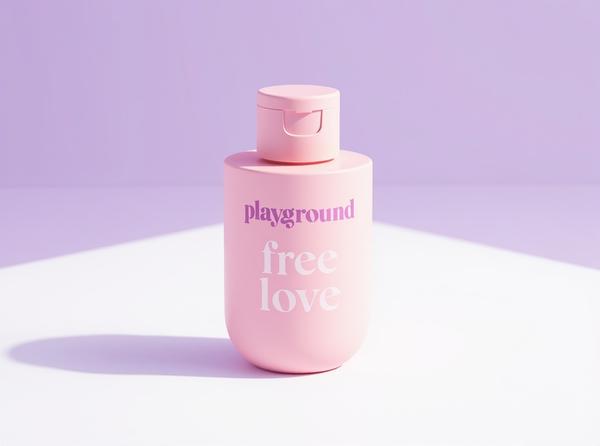 A pastel-pink Playground Free Love water-based lubricant bottle standing tall and bold on a bright plum-tinted studio backdrop, its label facing forward.