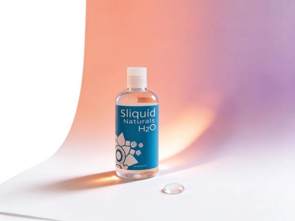 A clear bottle of Sliquid Naturals H2O water-based lubricant standing on a white paper surface in warm daylight, a soft coral and plum light wash on the wall behind it, with a single clear bead of lubricant resting on the paper beside its base.