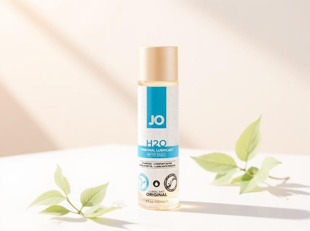 A bottle of JO H2O Original water-based lubricant standing upright in warm daylight on white paper with soft sage-green leaf sprigs and water droplets nearby, evoking gentle cleaning and safe care for silicone toys.