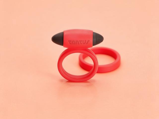 Crimson silicone Tantus vibrating cock ring standing upright with its removable bullet seated in the sleeve, and a second matching ring resting behind it on a warm peach paper background.