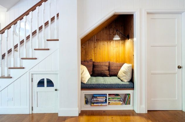 A small reading nook inset into the side of a white staircase with wood paneled walls, a green bench, white and brown throw pillows, and a small white overhead light 