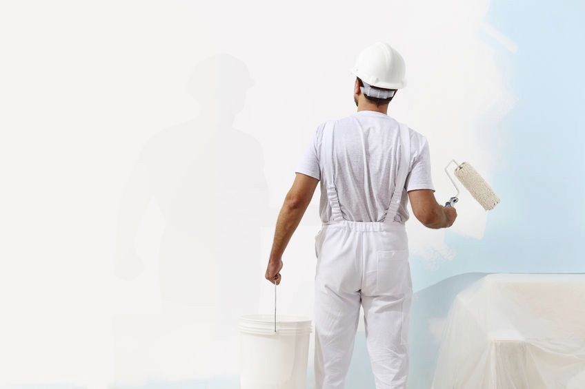 A man in a white painting uniform and white hard hat, with a paint roller in his right hand, facing a partially painted white wall