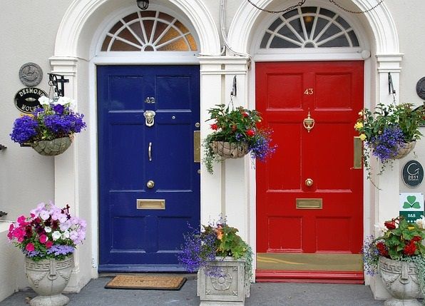 A blue front door to the left of a red front door, both with brass door knockers and colourful hanging plant baskets on either side